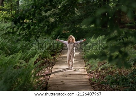 A little cute blonde girl runs along a wooden path among trees and ferns. Walking in the spring park, outdoor activities.Selective focus.