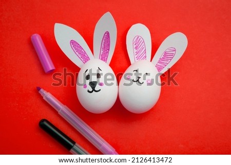 Photo of food for Easter. Chicken eggs with cute rabbit faces and bunny ears and purple and black felt-tip pens on red background. Easter coloring book for kids. Greeting card for Easter holiday.