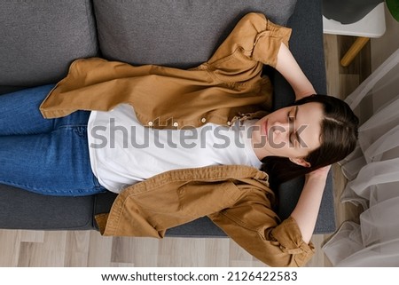 Top view of young woman enjoy day nap on comfy sofa. Caucasian female put hands behind hear lying on cushion on cozy grey couch breath fresh conditioned air inside modern living room. Repose concept Royalty-Free Stock Photo #2126422583