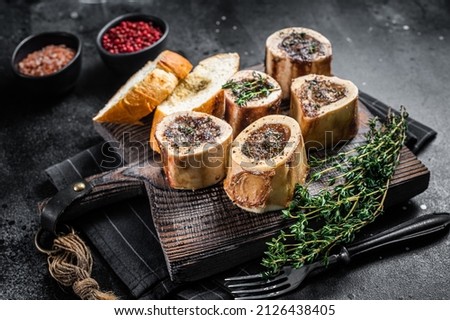 Roast marrow calf bones on wooden board with bread and herbs. Black background. Top view Royalty-Free Stock Photo #2126438405