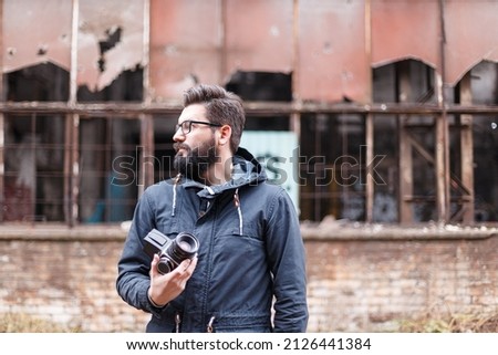 A bearded man with analog medium format camera , at damaged factory wall