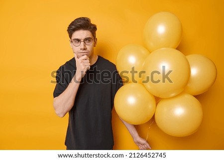 Serious thoughtful man concentrated at bunch of balloons thinks how to decorate hall for party wears round spectacles and casual black t shirt isolated over yellow background. Birthday celebration