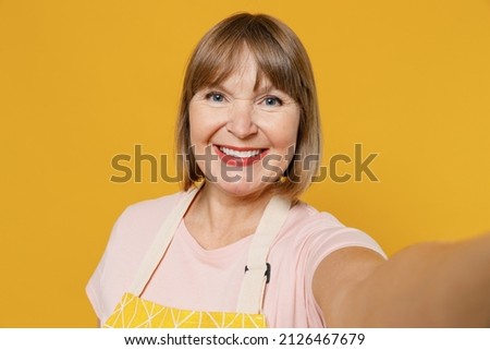 Close up smiling elderly housekeeper housewife woman 50s in orange apron doing selfie shot pov on mobile phone isolated plain on yellow background studio portrait. People household lifestyle concept