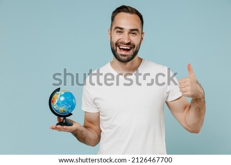 Young smiling happy fun geography teacher man wear blank print design white t-shirt hold in hands Earth world globe show thumb up gesture isolated on plain pastel light blue color background studio.
