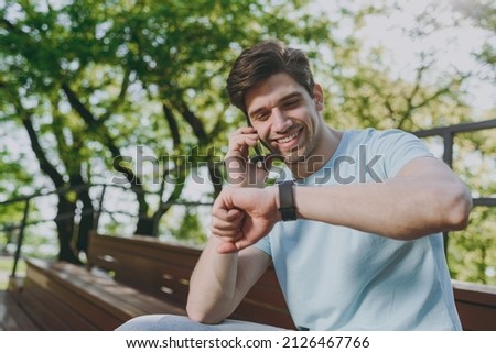 Smiling young happy man wear blue t-shirt sit on bench talk speak on mobile cell phone check time on smart watch rest relax in sunshine spring green city park outdoor on nature Urban leisure concept. Royalty-Free Stock Photo #2126467766