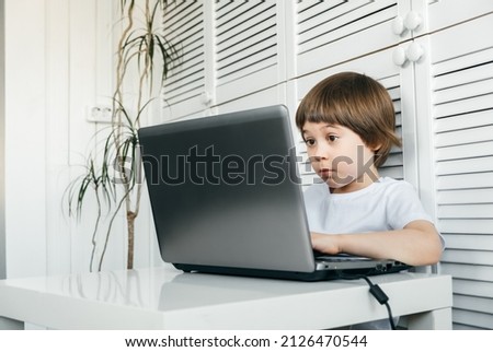 5 year boy sit at the table, uses laptop, looking at the screen. Child is doing homework lesson, playing video games, studying IT development, watching cartoon. Home distance online education concept.
