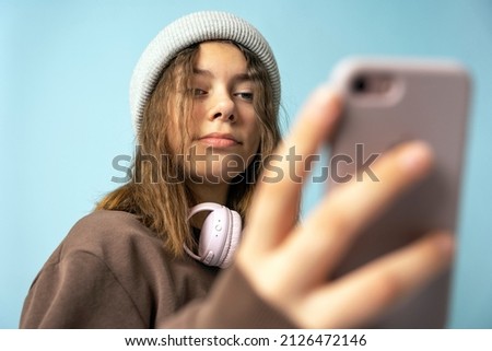A cute European teenage girl in fashionable knitted hat uses a smartphone and wireless headphones
