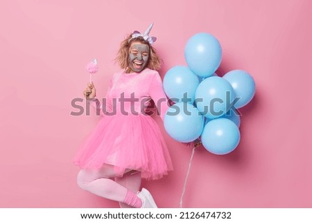 Overjoyed woman applies clay mask on face for rejuvenation dances carefree and sings has good mood during holiday wears unicorn hedband and dress holds bunch of blue helium balloons. Celebration