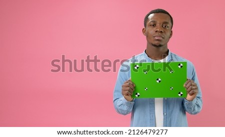 Serious African-American holding Banner with Green Screen Tracks Points for Copy Space. Empty Green Screen Board. Does Not Smile Looks at Camera With Space for Text or Advertising on Pink Background.