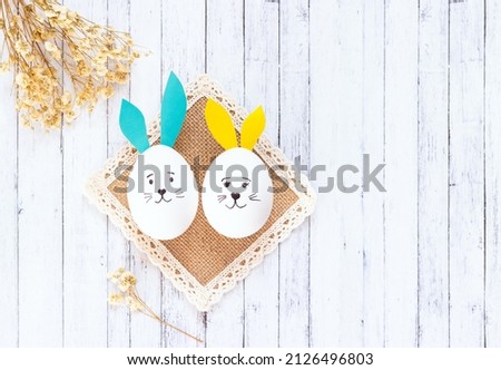 two white eggs with yellow and blue ears and a painted hare's face lie on a woven napkin. Funny Easter decor on a white wooden background, dried flowers. Copy space, top view