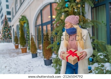 girl with a gift in red jacket and hat standing near a large Christmas tree.