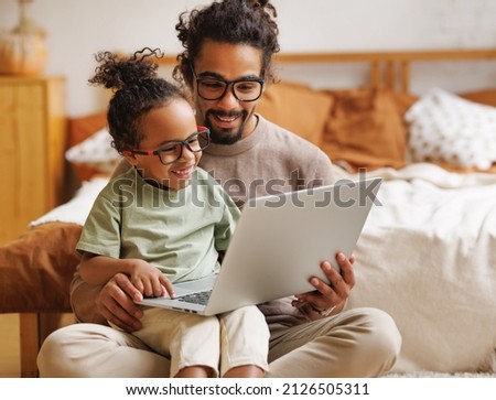 Happy african american family little boy son with young dad watching funny videos on laptop together at home, father with child using computer in bedroom while enjoying weekend