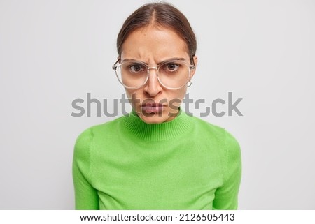 Serious dark haired woman looks with angry strict expression gazes directly at camera wears casual green jumper and big glasses for sight isolated over white background. Negative emotions concept Royalty-Free Stock Photo #2126505443