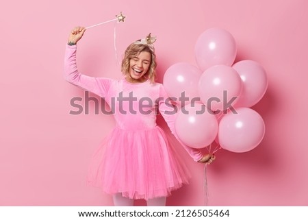 Overjoyed happy woman with hairstyle wears festive dress celebrates special occasion holds magical wand and bunch of helium balloons isolated over pink background. People and holidays concept