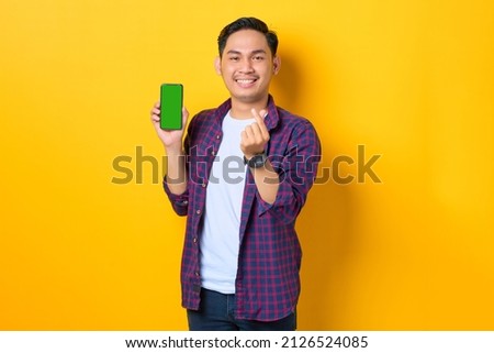 Smiling young Asian man in plaid shirt showing blank screen smartphone and gesturing korean heart with two fingers crossed isolated on yellow background