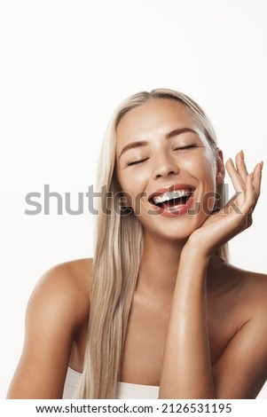 Vertical shot of beautiful blond woman model, laughing and smiling with white teeth, touching clear, clean glowing skin after skincare product, head and shoulders photo