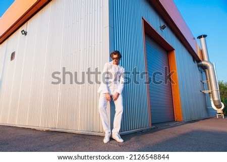 Wide angle portrait of a handsome man dressed in a white suit and round hippie sunglasses standing near the corner of a building during sunset