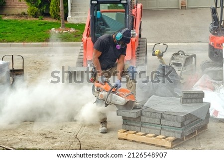 Landscaping worker wearing dust mask and cutting paving stones at residential driveway interlock pavement construction site.  Royalty-Free Stock Photo #2126548907