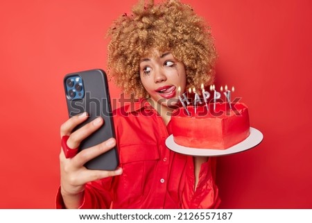 Funny woman with curly bushy hair sticks out tongue makes selfie via cellphone sticks out tongue holds heart cake celebrates anniversary wears shirt poses against red background. Celebration concept