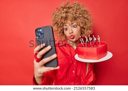 Curly haired young woman keeps lips folded makes selfie via smartphone poses with heart shaped cake going to celebrate specical occasion has spoiled makeup isolated over vivid red background.