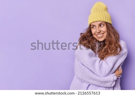 Tender pretty teenage girl with long hair embraces herself with love wears hat and warm winter jacket looks away gladfully isolated over purple background blank space for your advertising content