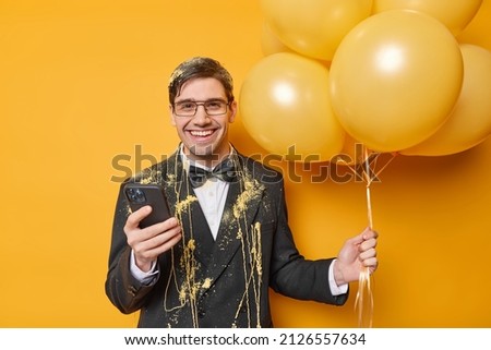 Positive man in festive clothes celebrates anniversary holds bunch of helium balloons and smartphone wears festive clothes being in good mood gets congratulations isolated over yellow background Royalty-Free Stock Photo #2126557634