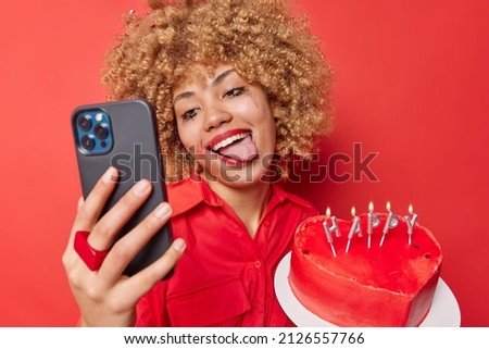 Positive carefree young woman sticks out tongue poses at smartphone camera for making selfie has festive mood holds cake in form of heart celebrates Valentines Day isolated over red background.