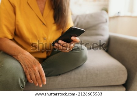Close-up picture of hands of elderly woman holding smartphone sitting on grey couch in yellow blouse and olive green jeans, wearing silver ring on finger, using gadget to surf Internet