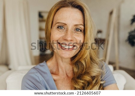 Senior woman-blogger with long golden hair, pretty freckled face, blue eyes and charming smile making selfie or recording stories for social network page, looking friendly and happy Royalty-Free Stock Photo #2126605607