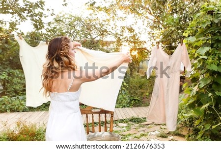 Young woman hanging laundry outdoors. Cute girl in dress washing white clothes in metal basin in backyard, hanging laundry on clothesline and leaving it to dry in garden, drying clothes outdoors  Royalty-Free Stock Photo #2126676353