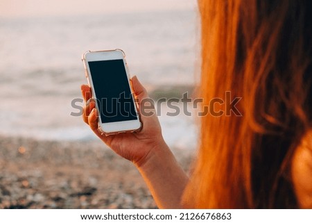 Unrecognizable young woman holding mobile phone at the beach against waves of sea. Sunset. Social media, modern technologies. Summertime vacations. Blogging