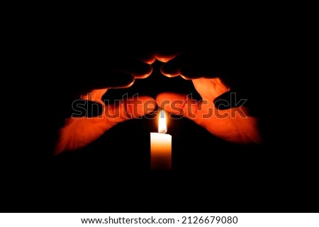 Hands are illuminated from the flame of a candle on a black background. The concept is the preservation of peace, warmth. ritual in magic. memory ritual. Royalty-Free Stock Photo #2126679080