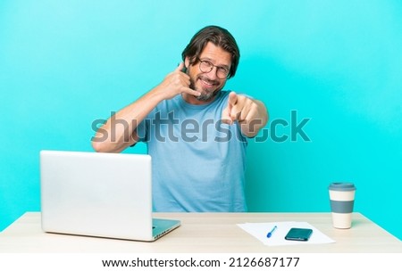 Senior dutch man in a table with a laptop isolated on blue background making phone gesture and pointing front