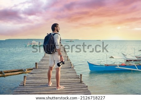 Photography and travel. Young man with rucksack holding camera standing on wooden fishing pier enjoying beautiful tropical sea view. Royalty-Free Stock Photo #2126718227