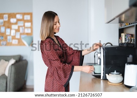 Woman using coffee machine to make big mug of coffee at home. Woman wearing silk robe at home while preparing a latte Royalty-Free Stock Photo #2126726507