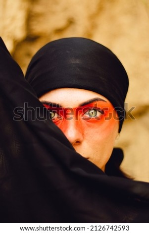 Portrait of a young beautiful woman with makeup and face art in American tribal or Middle Eastern style. Ethnic image, red eyes and black headscarf. Close-up.