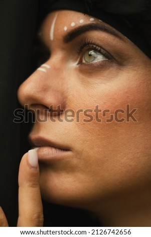 Portrait of a young beautiful woman with makeup and white face art ornament in tribal or middle eastern style dressed in a black hijab. Ethnic image Close-up on the background of a sandy wall.