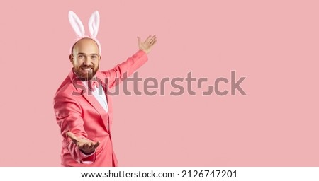 Funny man wearing bunny ears inviting you to Easter party. Happy young guy in pink suit and rabbit ears smiling and showing something on the right side on blank empty solid pink copy space background Royalty-Free Stock Photo #2126747201
