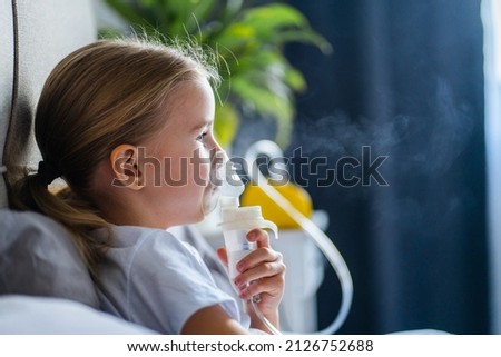 Sick little girl making inhalation with nebulizer to reduce coughing, lying in bed at home, child taking medication while breathing in through face mask. Bronchitis and asthma treatments for children Royalty-Free Stock Photo #2126752688