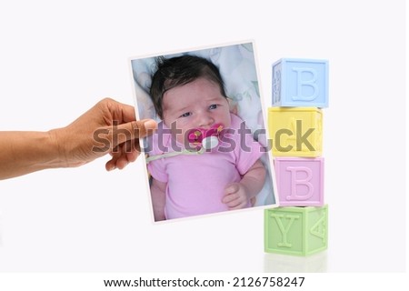Hand holding photograph of newborn infant baby girl next to stack of toy letter blocks on white background