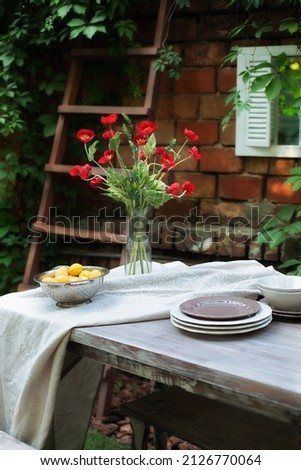 Summer bouquet of red poppies in a glass vase on Terrace. Spring Flowers in a vase at home. Wooden table and plates for garden party or dinner. Table for lunch outside in garden in patio yard of house