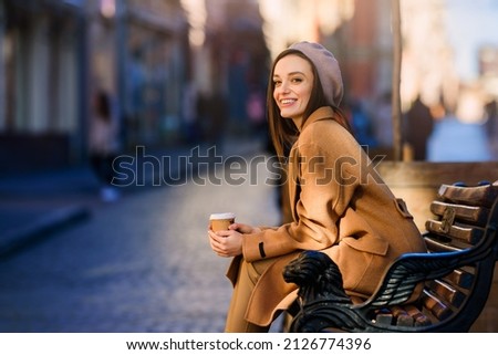 Young smiling woman girl drinking morning coffee resting on bench. Old town street background. Warm up with coffee in cold weather. Royalty-Free Stock Photo #2126774396