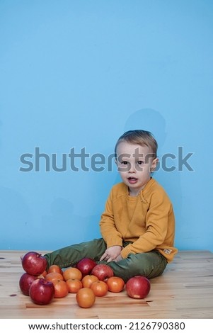 2 year old boy with red apples and tangerines. boy dressed green pants yellow jacket sits advertising vegetables fruits