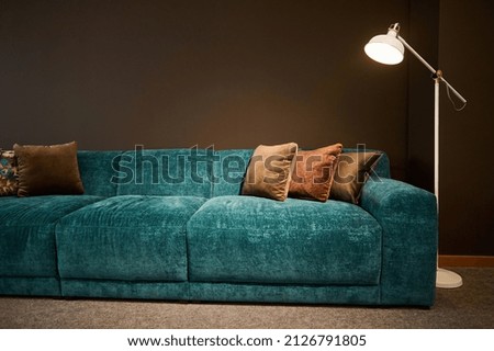 Exhibition of modern stylish upholstered furniture in the showroom of a furniture store. Focus on a turquoise soft velour sofa and brown pillows lit by a lamp against a brown wall background Royalty-Free Stock Photo #2126791805