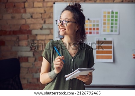 Smiling woman, interior designer or architect in casual wear with messy hairdo holding pen and notebook, looking aside, working on new creative project, standing near board with color samples Royalty-Free Stock Photo #2126792465