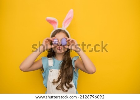 A cute girl with dark long hair in a headband with rabbit ears holds purple Easter eggs in her hands in front of her eyes and kisses. Studio portrait on a yellow background. Happy childhood, happy
