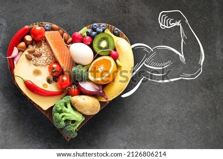 A healthy, balanced diet. Healthy food on a heart-shaped wooden cutting board and a muscular arm as a synonym for strength and health. Royalty-Free Stock Photo #2126806214