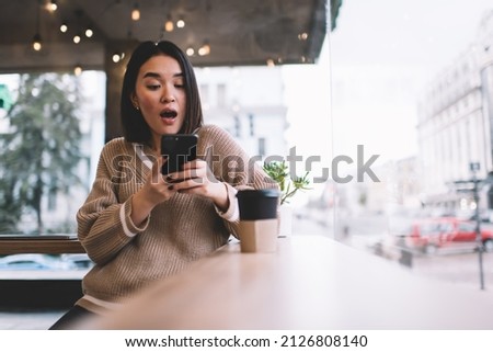 Surprised asian girl using and watching on smartphone in cafe. Young millennial brunette woman standing at table near window. Concept of rest, leisure and free time. Modern female lifestyle