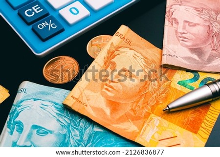 Brazilian Real banknotes on a dark wooden table with a calculator, a pen and some coins in the image composition. Concepts of Brazilian economy and finance.
