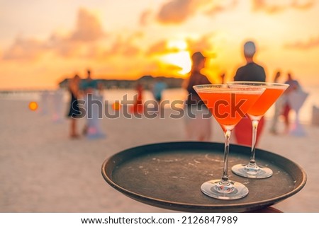 Two cocktail drinks with blur beach party people and colorful sunset sky in the background. Luxury outdoor leisure lifestyle, relaxing and romantic colors, blurred people partying on a summer evening